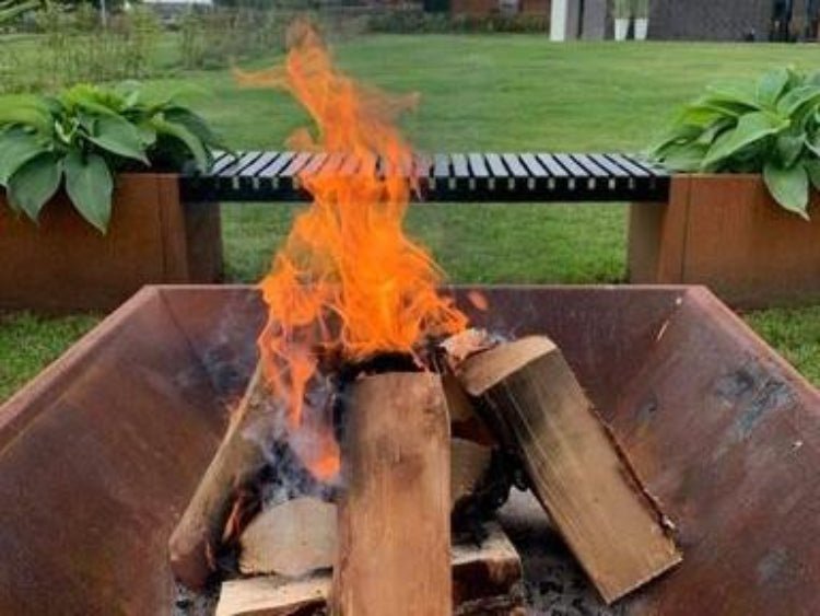 Firkantet bålplass i cortenstål med ved og levende flammer, plassert i hage