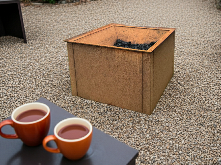 Firkantet bålplass i cortenstål plassert på grus, med kull og hagemiljø rundt