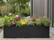 Lang plantekasse i svart stål med blomstrende planter i hage